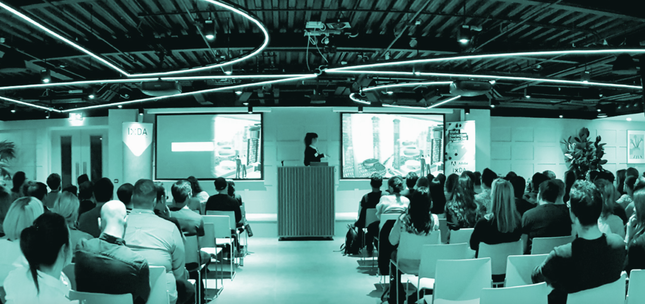 A woman behind a lectern at the front of a classroom. Two screens are on either side of her.